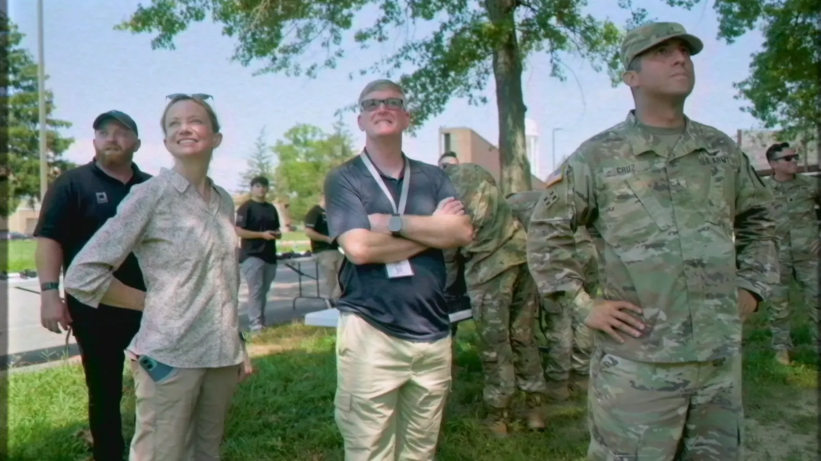 Soldiers and civilians gathered outdoors at a demonstration, smiling as they watch a flight