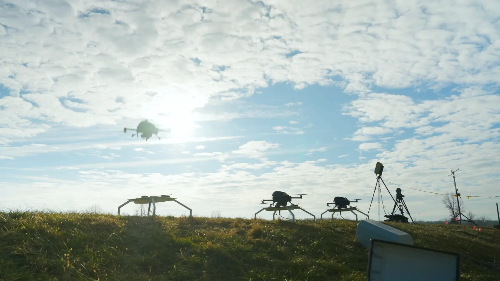 Drones staged on a grass line against a bright sky, one in flight