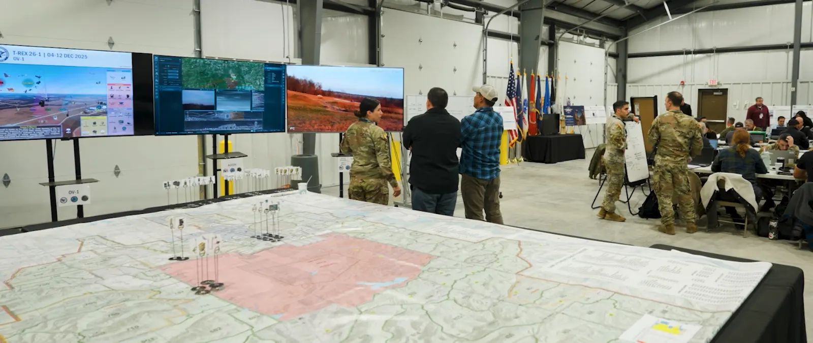 Service members and civilians at a brief in a hangar, a large terrain map on the floor